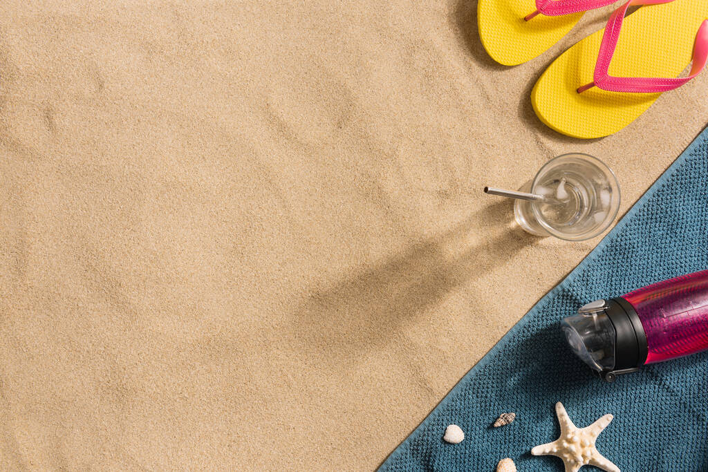 夏休みの構成。砂の背景に水のフリップフロップ、水ボトルとガラス。影の強い光。夏の背景。タオル製のボーダー構成 - 写真・画像