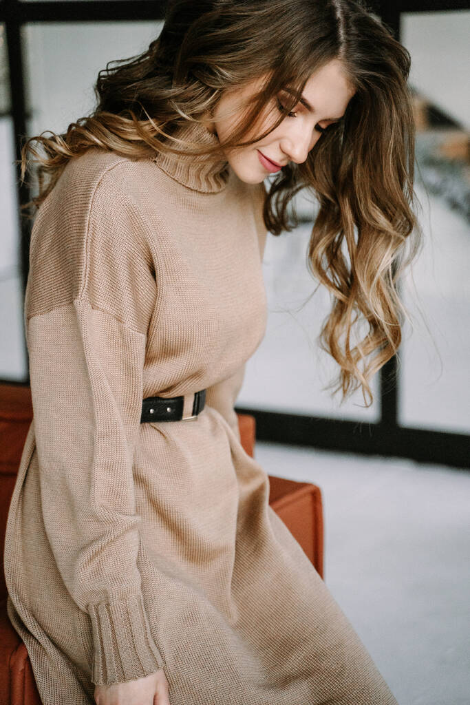 Ξανθιά κοπέλα με μακριά μαλλιά σε ένα καφέ πλεκτό φόρεμα σε ένα κομψό διαμέρισμα - Φωτογραφία, εικόνα