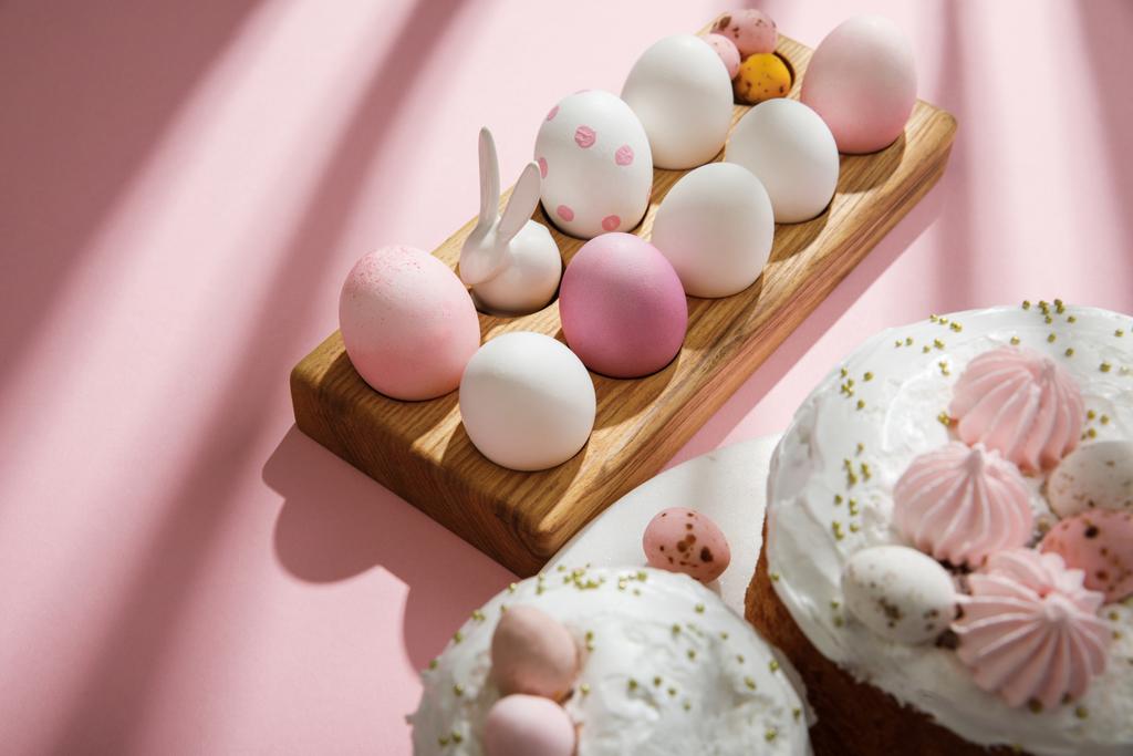 Enfoque selectivo de huevos pintados de pollo y codorniz, conejito decorativo en tablero de madera cerca de pasteles de Pascua sobre fondo rosa
 - Foto, Imagen