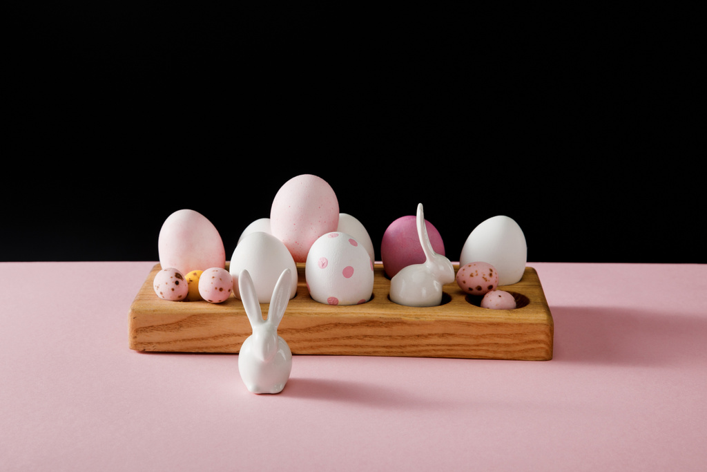 Coelhinhos brancos decorativos e ovos de páscoa em tábua de madeira sobre fundo rosa e preto
 - Foto, Imagem