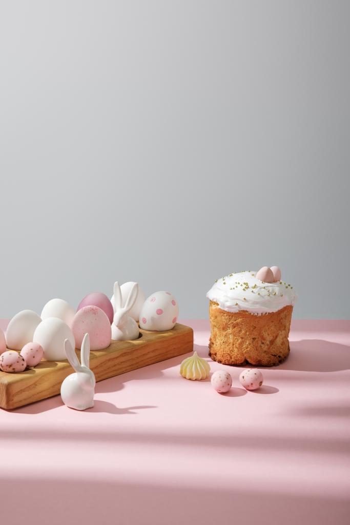 Пасхальные яйца на деревянной доске с декоративными кроликами возле пасхального торта на розовом и сером фоне
 - Фото, изображение