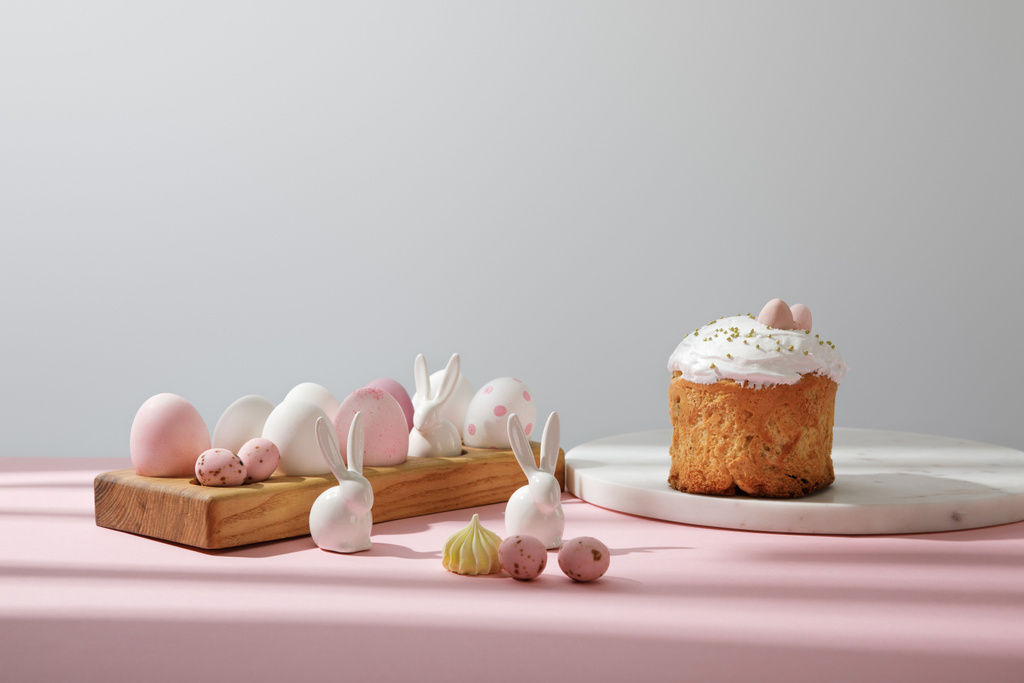 Пасхальные яйца на деревянной доске с декоративными кроликами возле пасхального хлеба на розовом и сером фоне
 - Фото, изображение