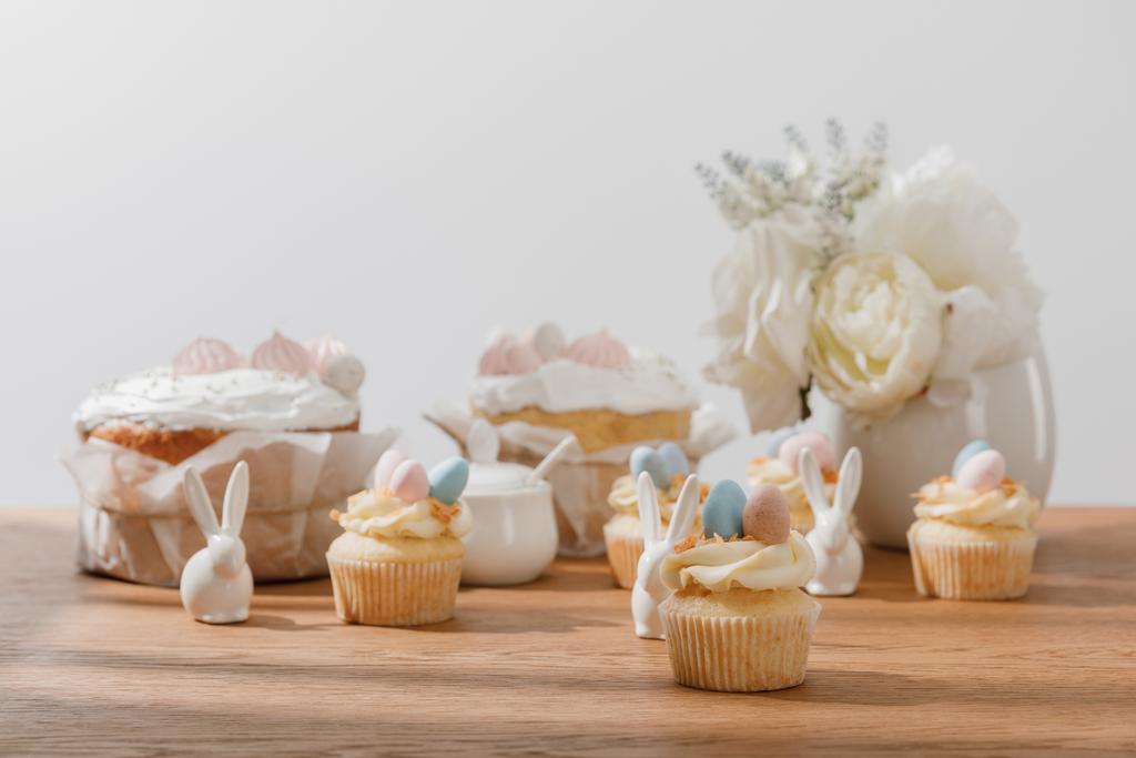 Selectieve focus van cupcakes met decoratieve konijntjes, suikerpot, paastaart en vaas met bloemen geïsoleerd op grijs - Foto, afbeelding