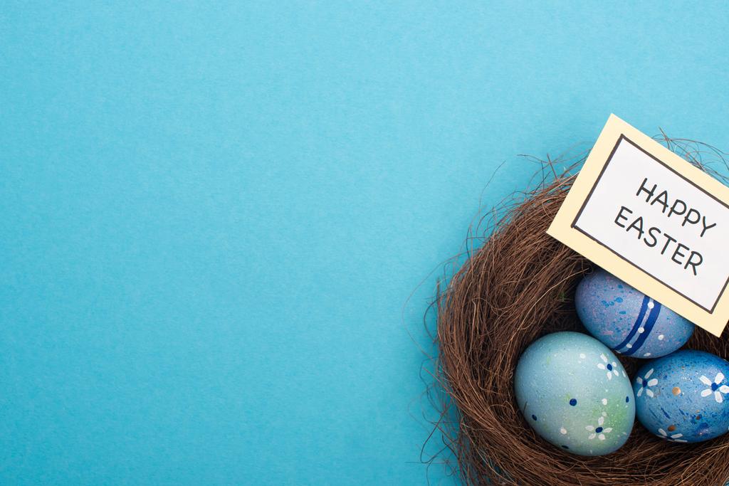 Vista superior del nido con huevos de Pascua y tarjeta con letras de Feliz Pascua sobre fondo azul
 - Foto, imagen