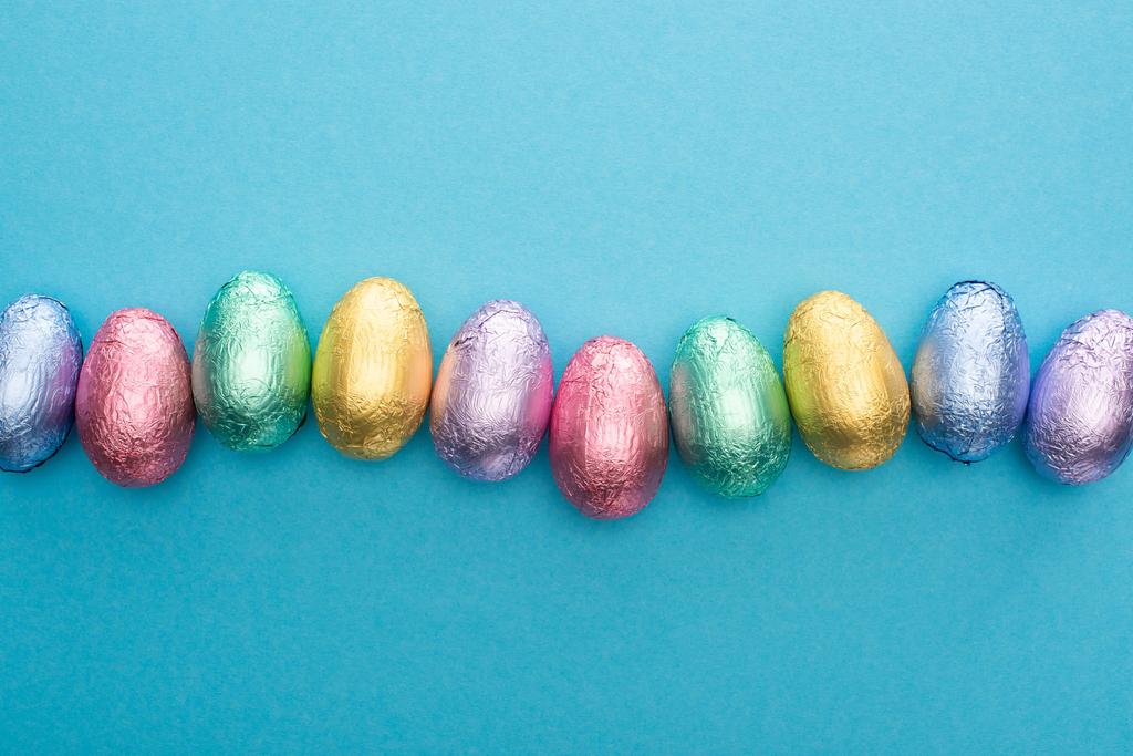 Верхний вид шоколадных пасхальных яиц в красочной фольге на голубом фоне
 - Фото, изображение
