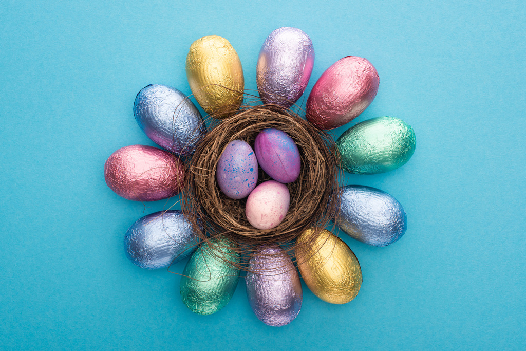 Bovenaanzicht van chocolade Paaseieren in kleurrijke folie rond nest met kwarteleitjes op blauwe achtergrond - Foto, afbeelding