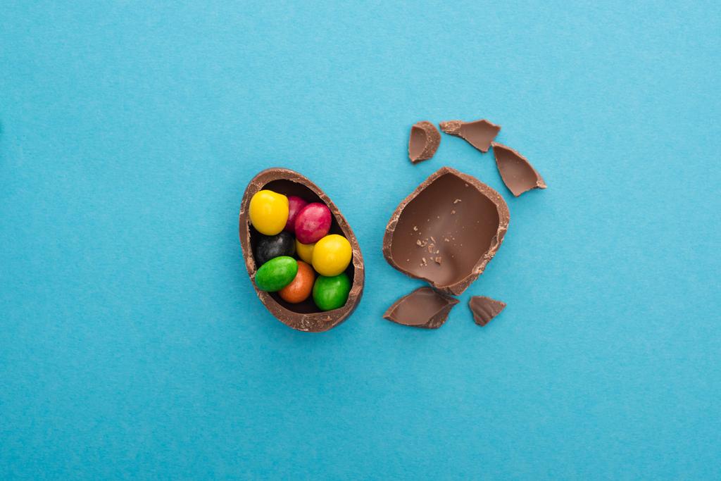 Ansicht von Stücken und der Hälfte der Schokolade Osterei mit bunten Bonbons auf blauem Hintergrund - Foto, Bild
