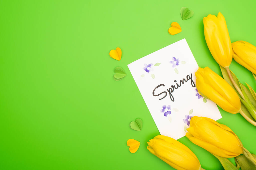 Widok z góry żółte tulipany, kartka z wiosennym napisem i dekoracyjne serca na zielonym tle - Zdjęcie, obraz