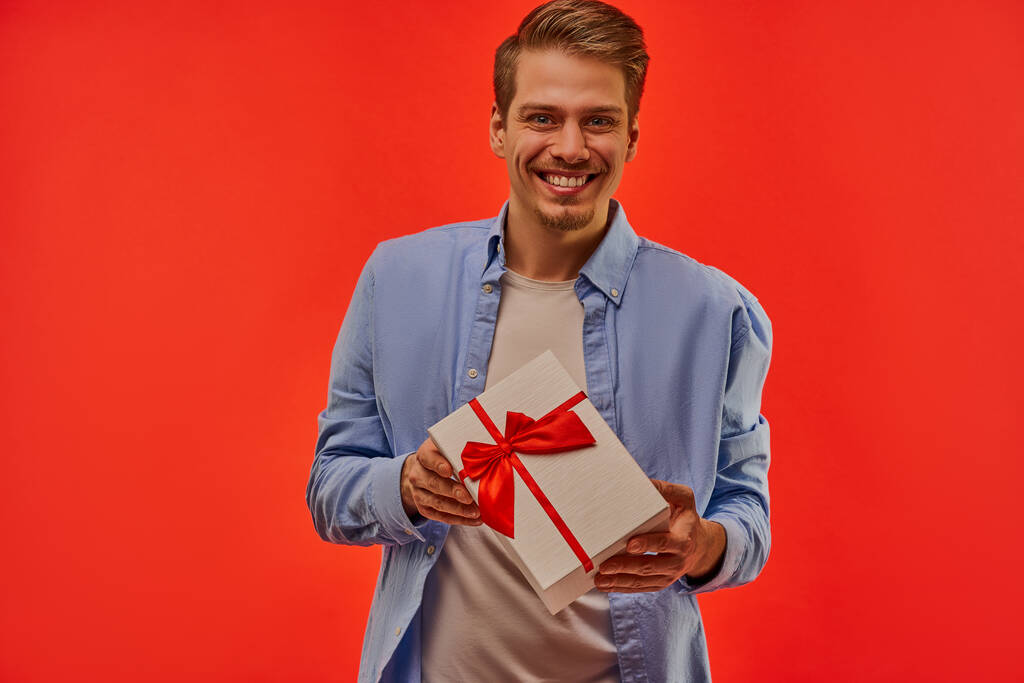 Un tipo sonríe ancho con caja blanca con un lazo rojo y cintas en las manos
. - Foto, Imagen