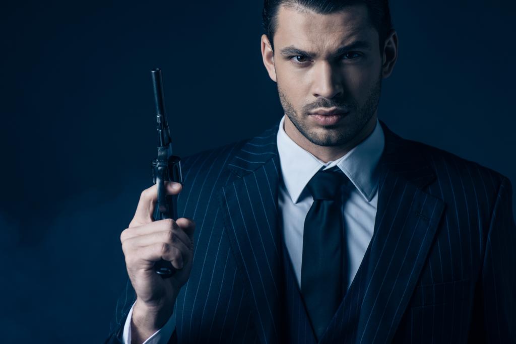 Vista frontal do gangster segurando arma e olhando para a câmera no fundo azul escuro
 - Foto, Imagem