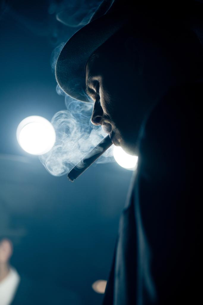 Vista de ángulo bajo del cigarro fumador mafioso en la oscuridad
 - Foto, imagen