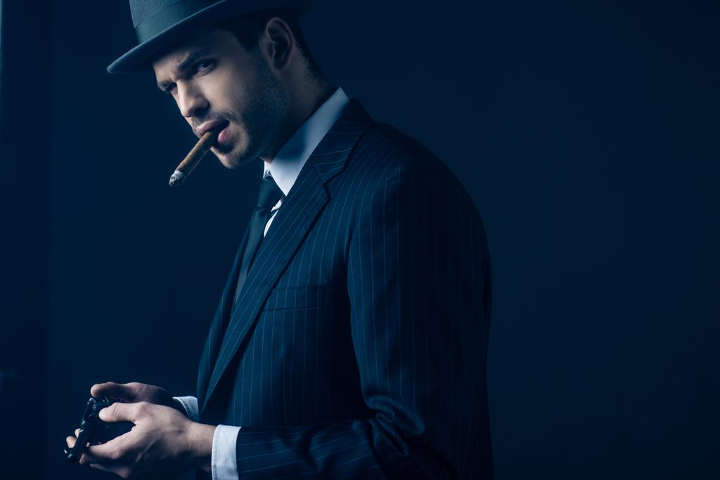 Gángster con cigarro en la boca pistola de carga sobre fondo azul oscuro
 - Foto, Imagen