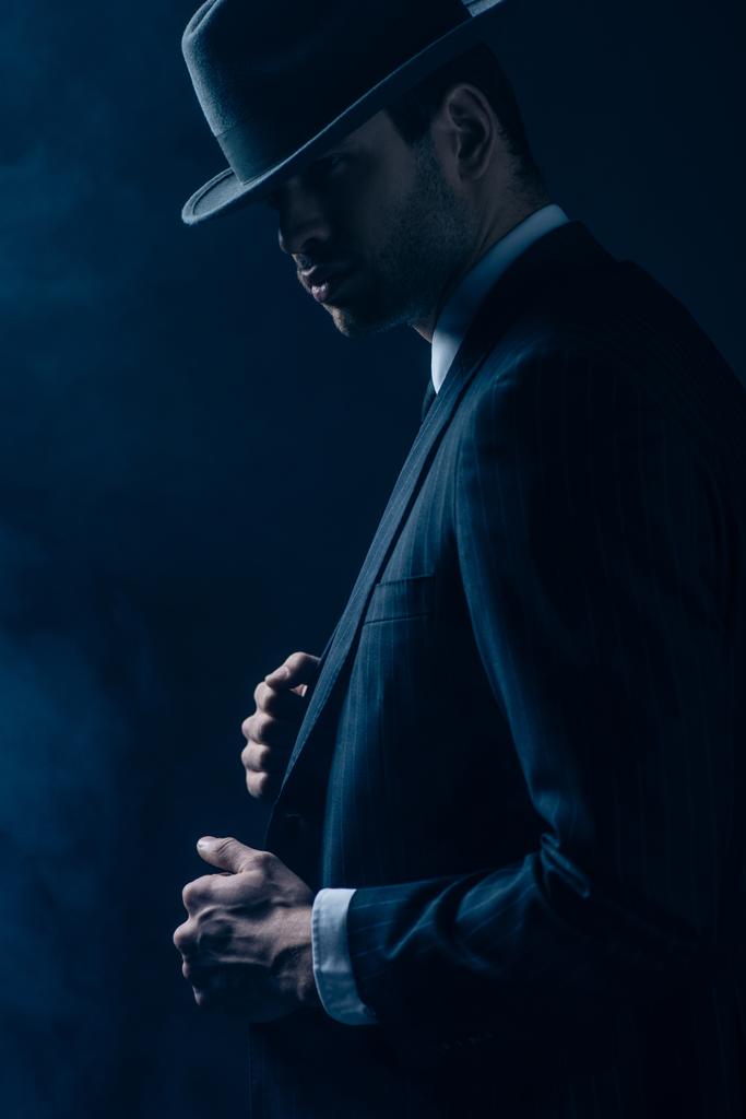 Vista lateral de mafioso confiante em terno preto e chapéu de feltro no fundo escuro
 - Foto, Imagem