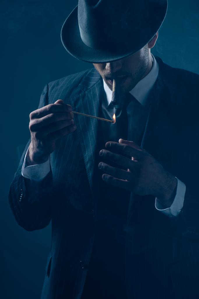 Mafioso with covered eyes with felt hat lighting cigar on dark blue  - Photo, Image