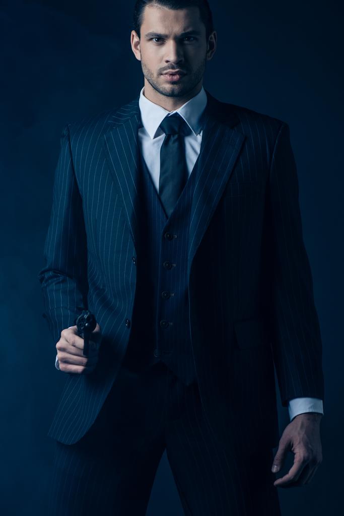 Vista frontal de un gángster confiado apuntando con el arma y mirando a la cámara sobre fondo azul oscuro
 - Foto, imagen