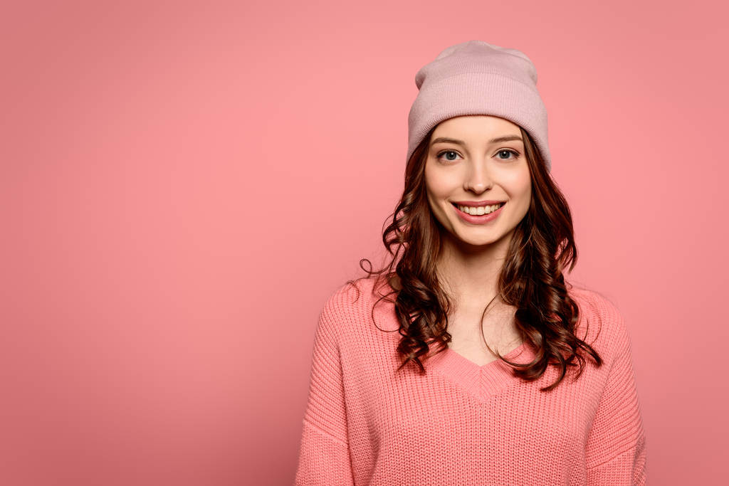 attractive cheerful girl in sweater and hat smiling at camera isolated on pink - Photo, Image