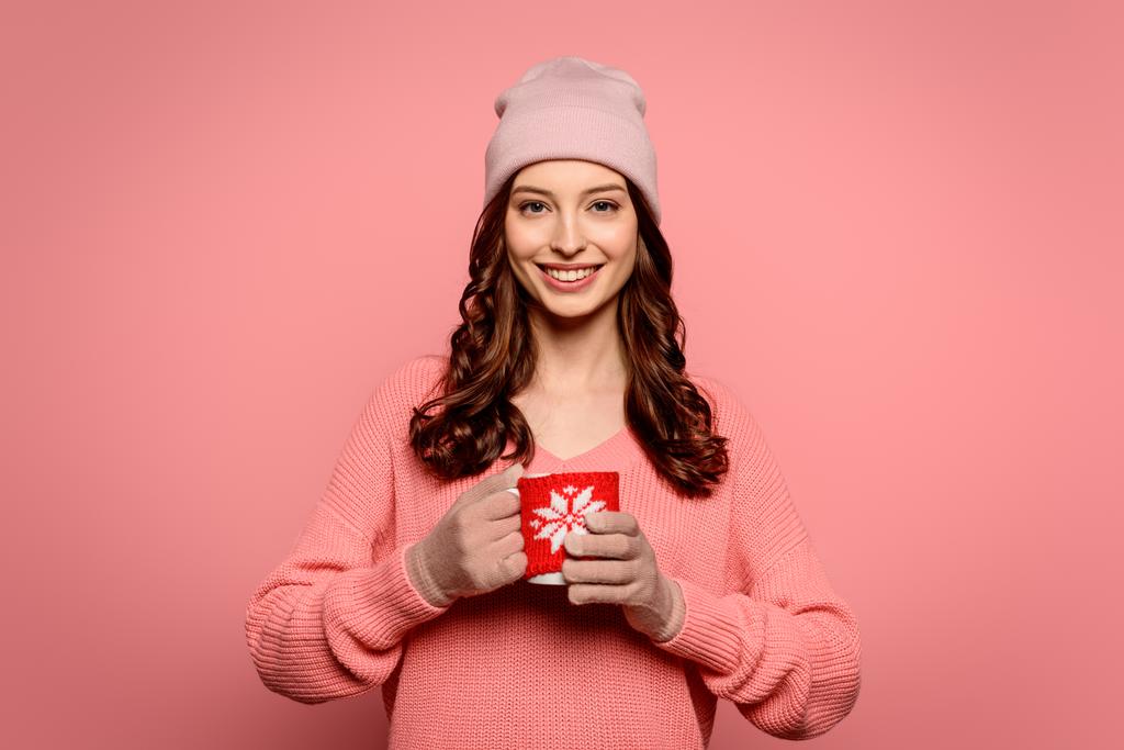 glimlachend meisje in hoed en handschoenen houden mok met warm drankje geïsoleerd op roze - Foto, afbeelding