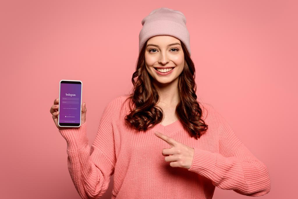 KYIV, UKRAINE - NOVEMBER 29, 2019: smiling girl pointing with finger at smartphone with Instagram app on screen isolated on pink - Fotó, kép