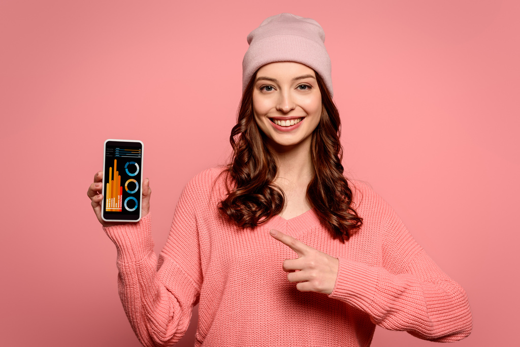 alegre chica apuntando con el dedo en el teléfono inteligente con gráficos y gráficos en la pantalla aislado en rosa
 - Foto, imagen