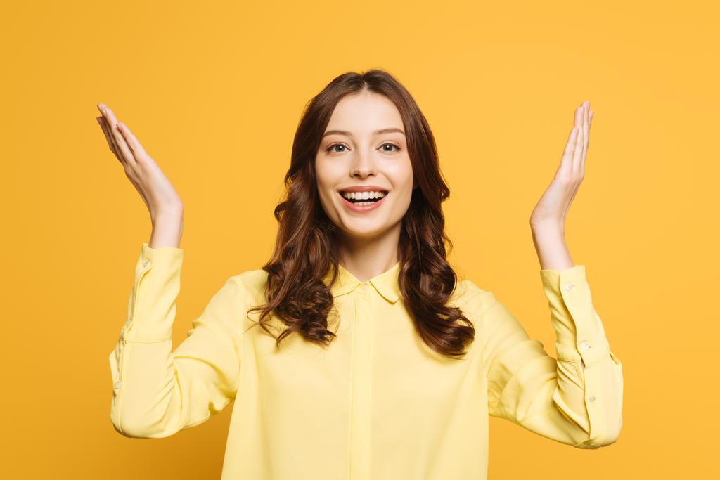 excited girl showing wow gesture with raised hands while smiling at camera isolated on yellow - Photo, Image