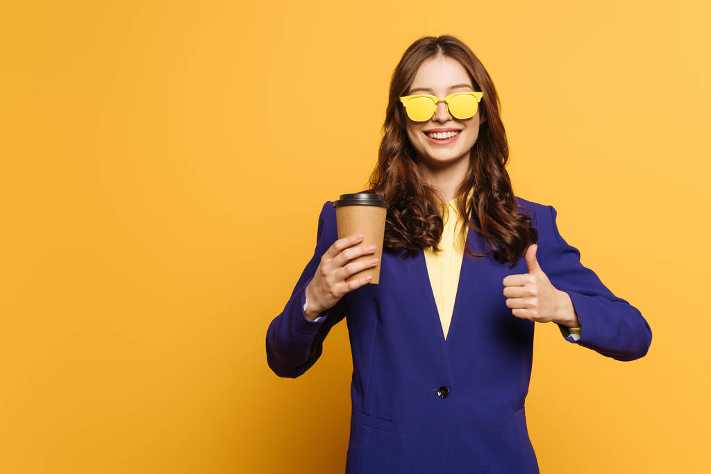 stylové, usmívající se dívka ve žlutých brýlích drží kávu jít a ukazující palec nahoru na žlutém pozadí - Fotografie, Obrázek