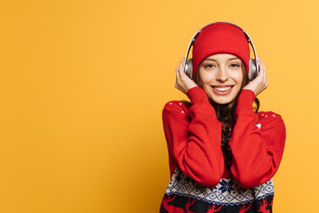 smiling girl in hat and red ornamental sweater listening music in wireless headphones on yellow background - Photo, Image