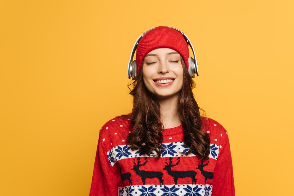 glückliches Mädchen mit Hut und rotem Zierpullover, das Musik in drahtlosen Kopfhörern hört, mit geschlossenen Augen, isoliert auf gelb - Foto, Bild