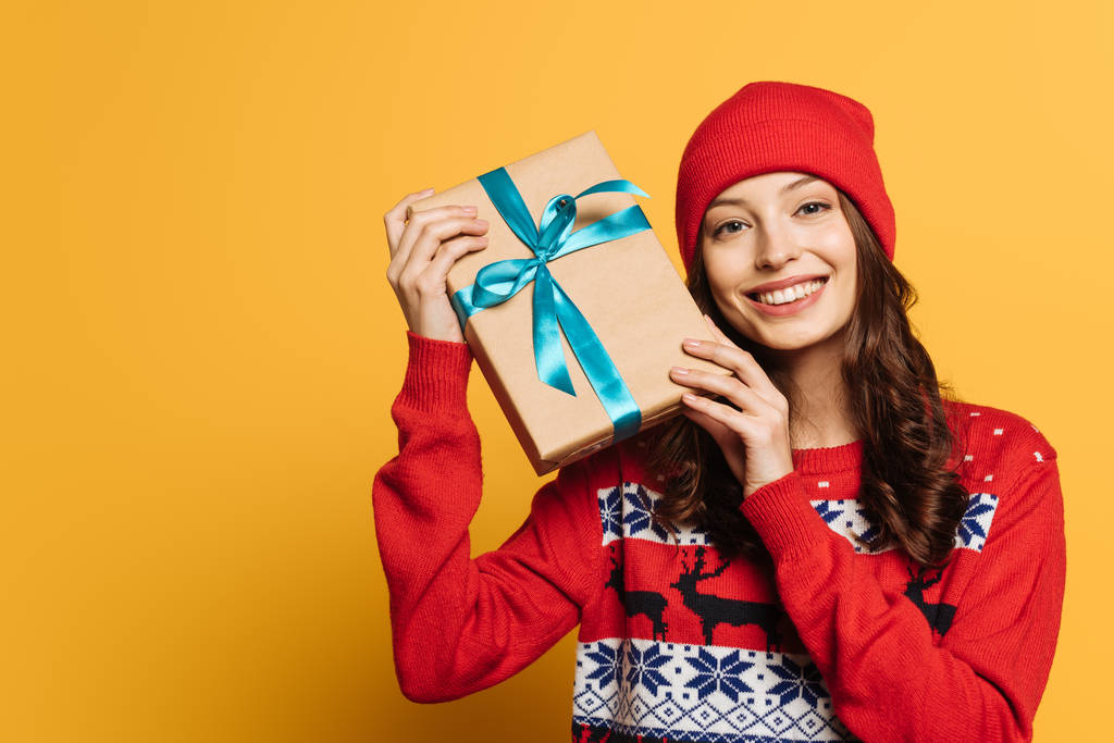 fröhliches Mädchen mit Hut und rotem Zierpullover mit Geschenkbox auf gelbem Hintergrund - Foto, Bild