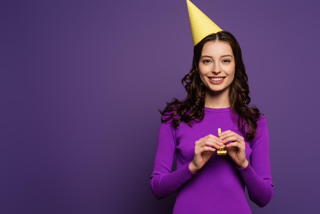 счастливая девушка в кепке, держащая партийный рог и улыбающаяся камере на фиолетовом фоне
 - Фото, изображение