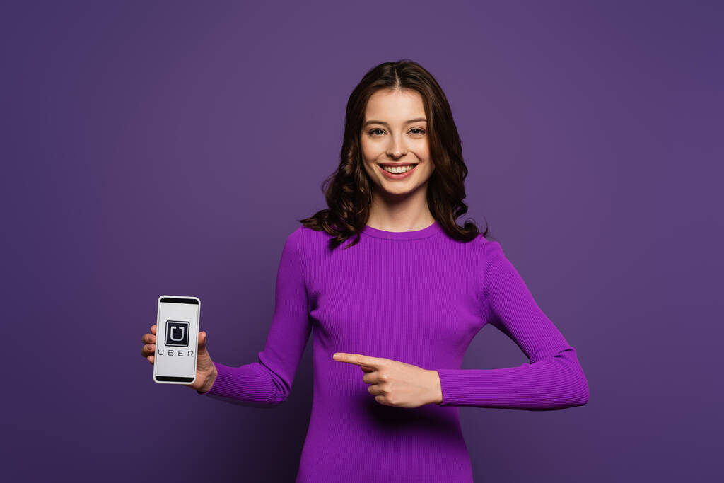KYIV, UKRAINE - NOVEMBER 29, 2019: smiling girl pointing with finger at smartphone with Uber app on screen on purple background - Photo, Image