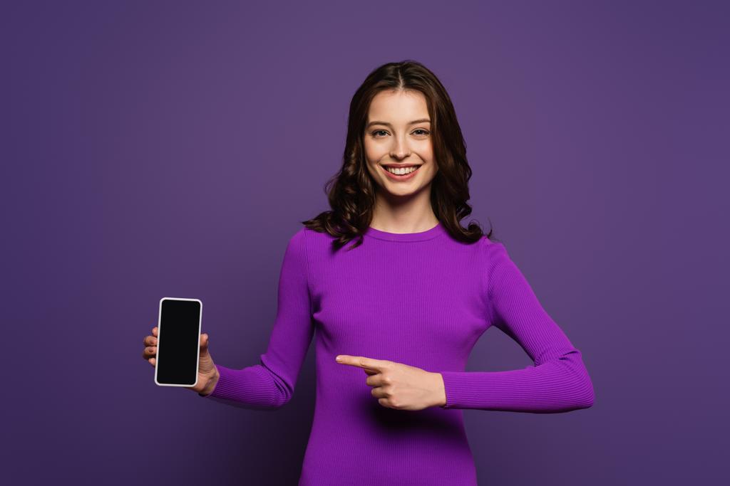 chica sonriente apuntando con el dedo al teléfono inteligente con pantalla en blanco sobre fondo púrpura
 - Foto, imagen