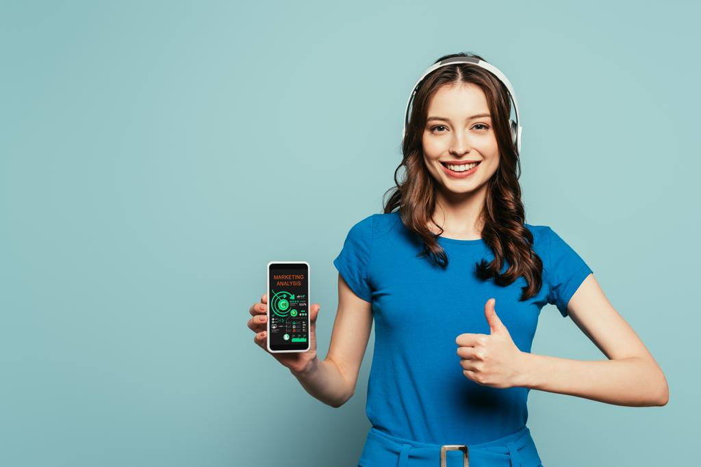 Kablosuz kulaklıklı mutlu kız mavi arka planda pazarlama analizi uygulaması ile akıllı telefonu tutarken baş parmağını kaldırıyor. - Fotoğraf, Görsel