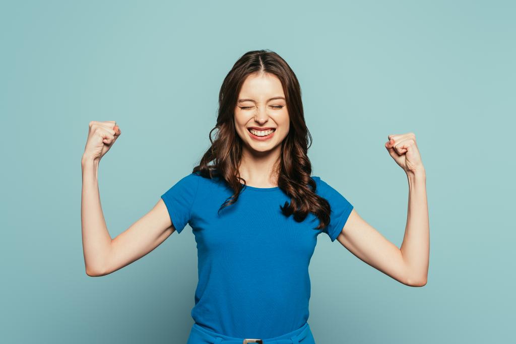 vzrušená dívka ukazuje vítězný gesto se zavřenýma očima při úsměvu na modrém pozadí - Fotografie, Obrázek