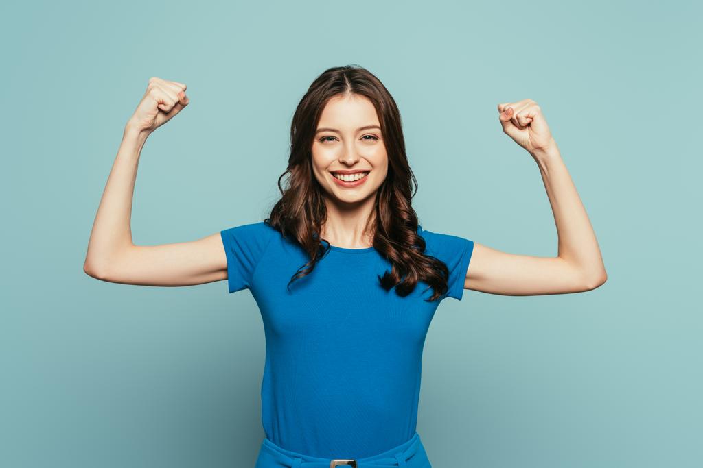 cheerful girl showing winner gesture while smiling at camera on blue background - Photo, Image