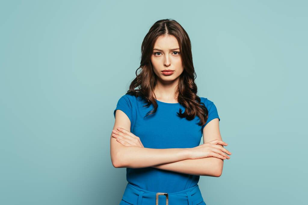 serious, attractive girl looking at camera while standing with crossed arms on blue background - Photo, Image