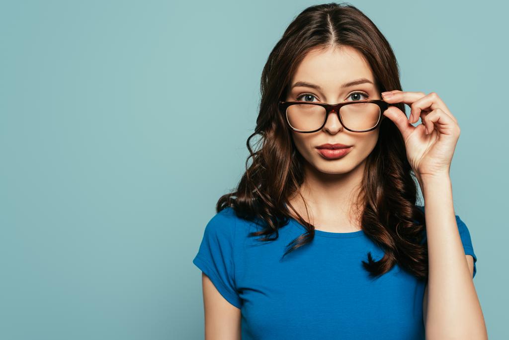 atractiva, chica segura de tocar gafas mientras mira la cámara aislada en azul
 - Foto, Imagen