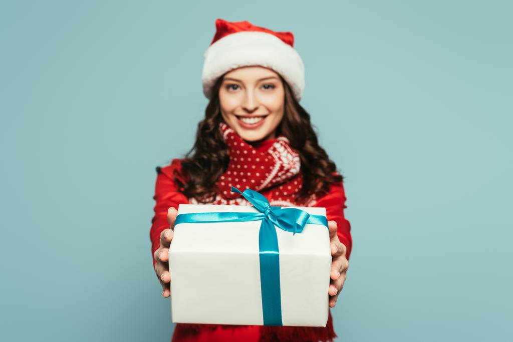 allegra ragazza in cappello di Babbo Natale che mostra scatola regalo mentre guarda la fotocamera su sfondo blu
 - Foto, immagini