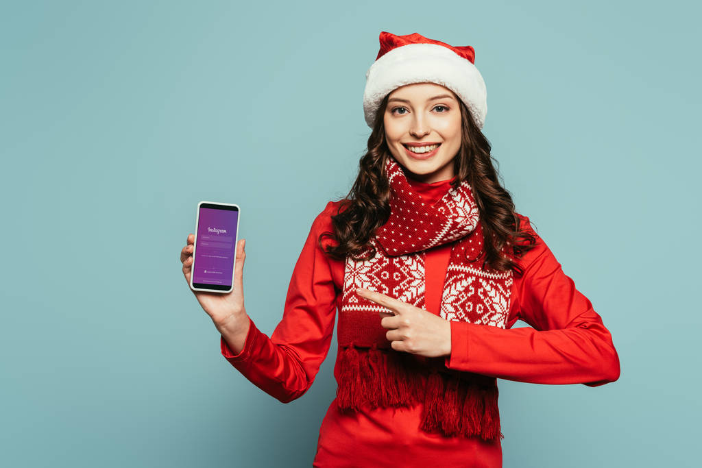 1kyiv,ウクライナ- 2019年11月29日:幸せな女の子でサンタの帽子と赤いセーターポインティングで指でスマートフォンでInstagramのアプリで画面上の青の背景 - 写真・画像