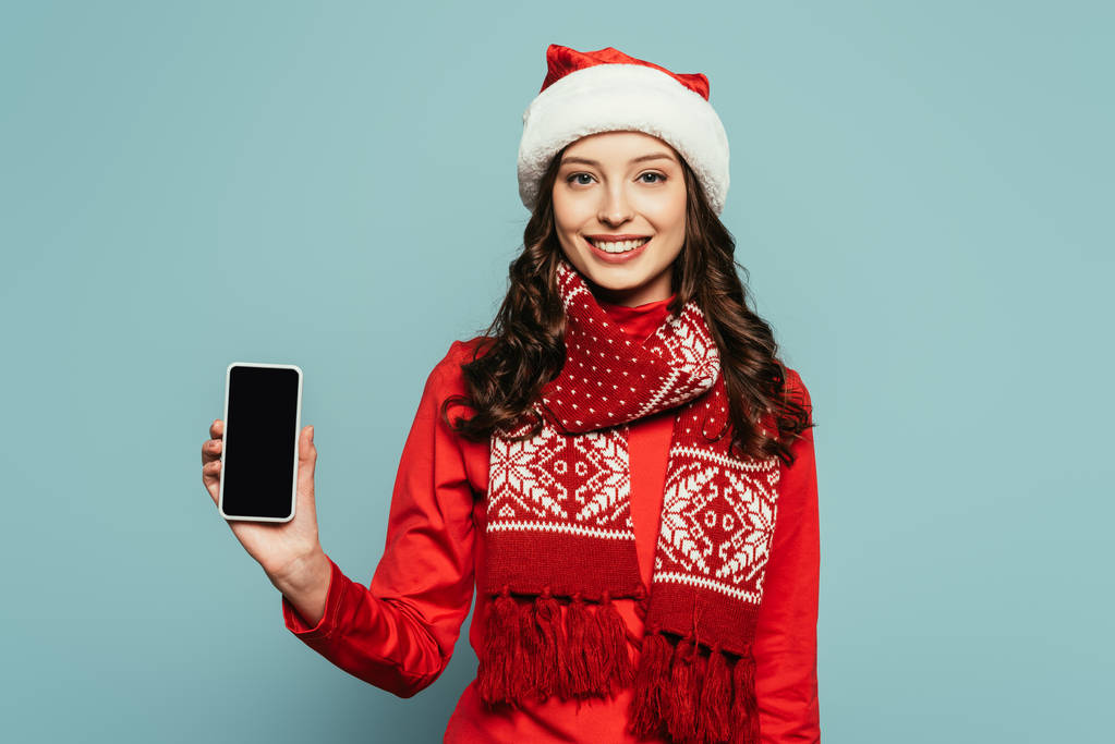 glimlachend meisje in santa hoed en rode trui tonen smartphone met leeg scherm op blauwe achtergrond - Foto, afbeelding