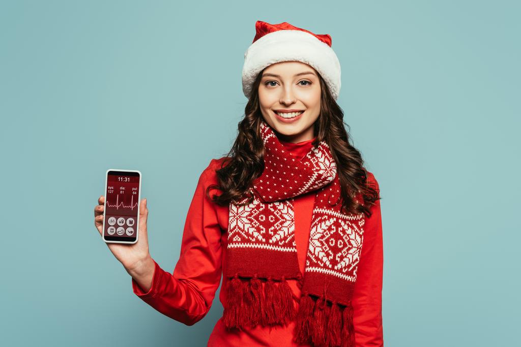 cheerful girl in santa hat and red sweater showing smartphone with heartbeat rate on screen on blue background - Photo, Image