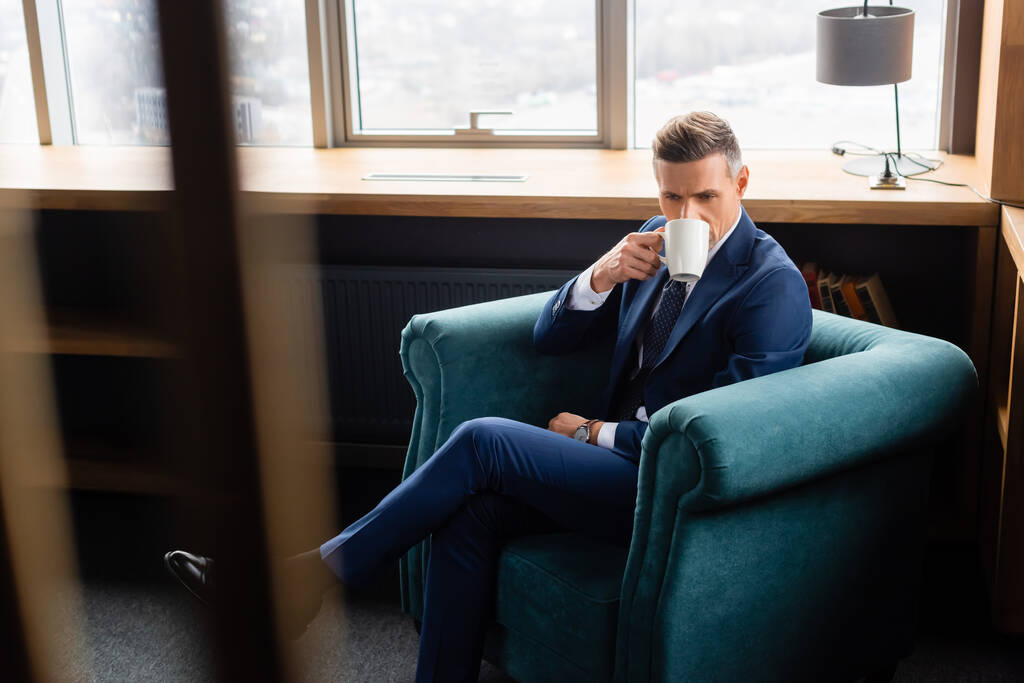 επιλεκτική εστίαση του επιχειρηματία στο κοστούμι πίνοντας καφέ και κάθεται στην πολυθρόνα  - Φωτογραφία, εικόνα