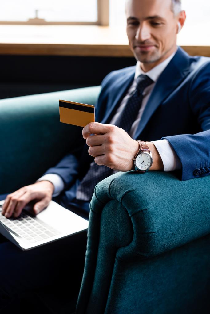 επιλεκτική εστίαση του χαμογελαστού επιχειρηματία με κοστούμι κρατώντας πιστωτική κάρτα και χρησιμοποιώντας φορητό υπολογιστή  - Φωτογραφία, εικόνα