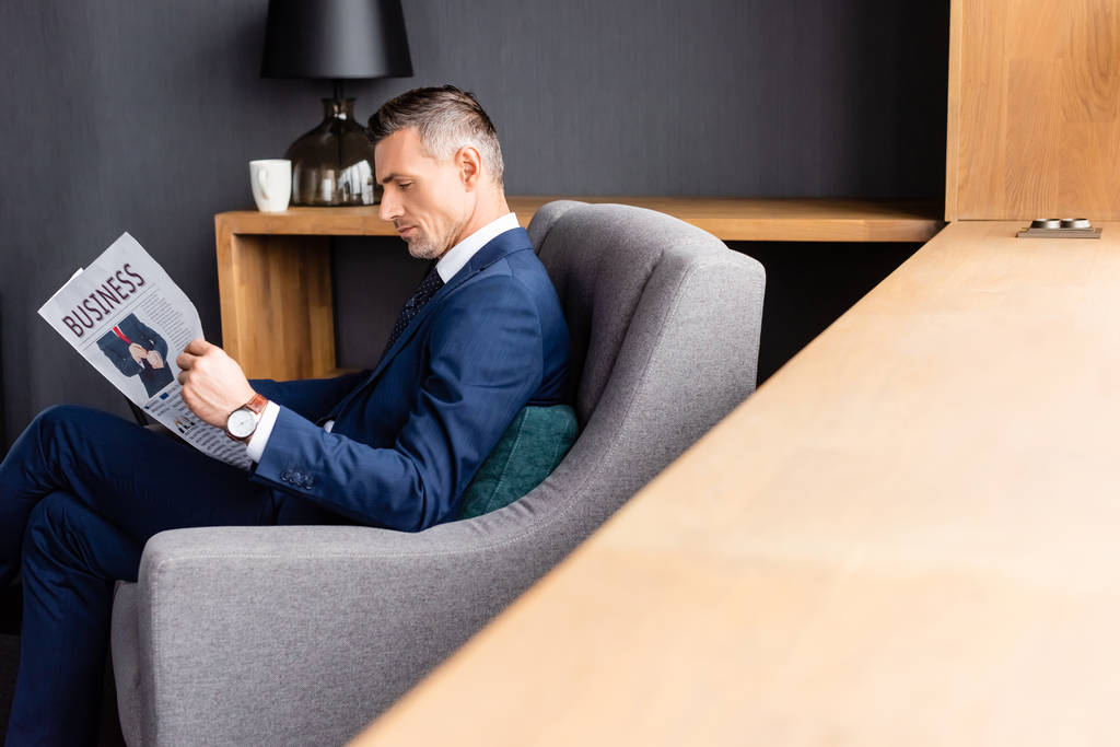 vista lateral del hombre de negocios en traje leyendo periódico con letras de negocios y sentado en sillón
  - Foto, Imagen