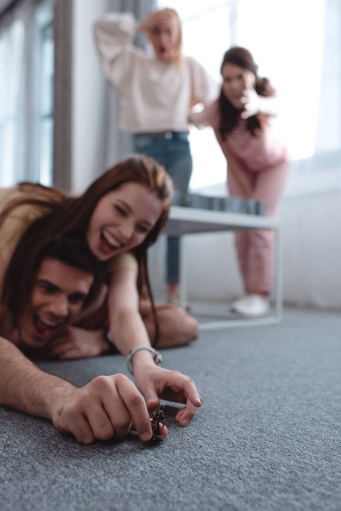 selective focus of cheerful girl trying to take toy figurine from guy lying on floor and excited girls standing on background - Photo, Image