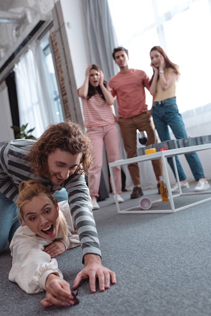 KYIV, UCRANIA - 27 de enero de 2020: enfoque selectivo del tipo que trata de tomar la figura de juguete de una chica acostada en el suelo cerca de amigos sorprendidos de pie cerca del juego de laberinto
 - Foto, Imagen