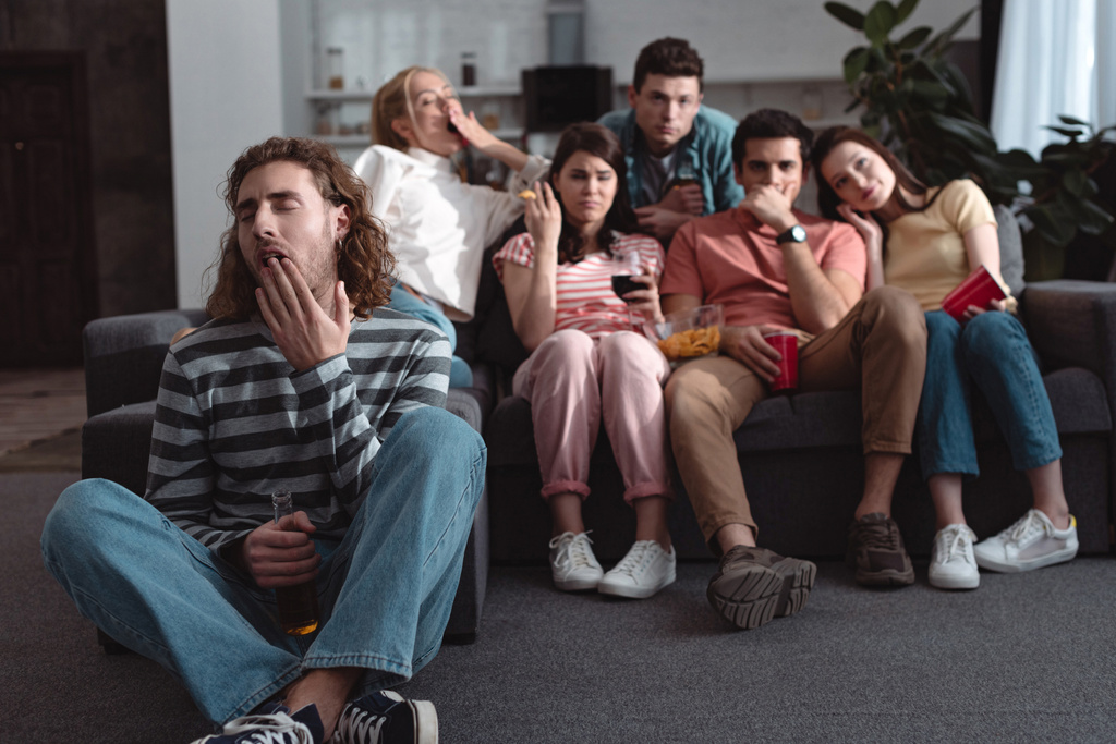 valikoiva painopiste nuori mies haukottelu istuessaan lattialla lähellä tylsää ystäviä katsomassa televisiota kotona
 - Valokuva, kuva