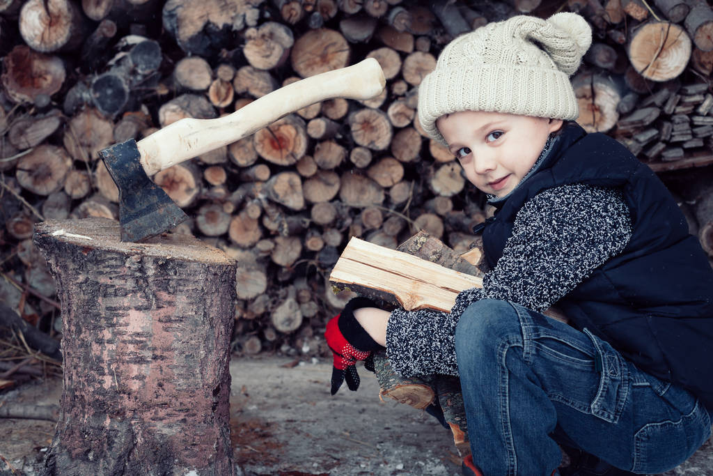Un petit garçon coupant du bois de chauffage dans la cour avant le jour. Le gamin s'amuse dehors. Concept mode de vie heureux
. - Photo, image