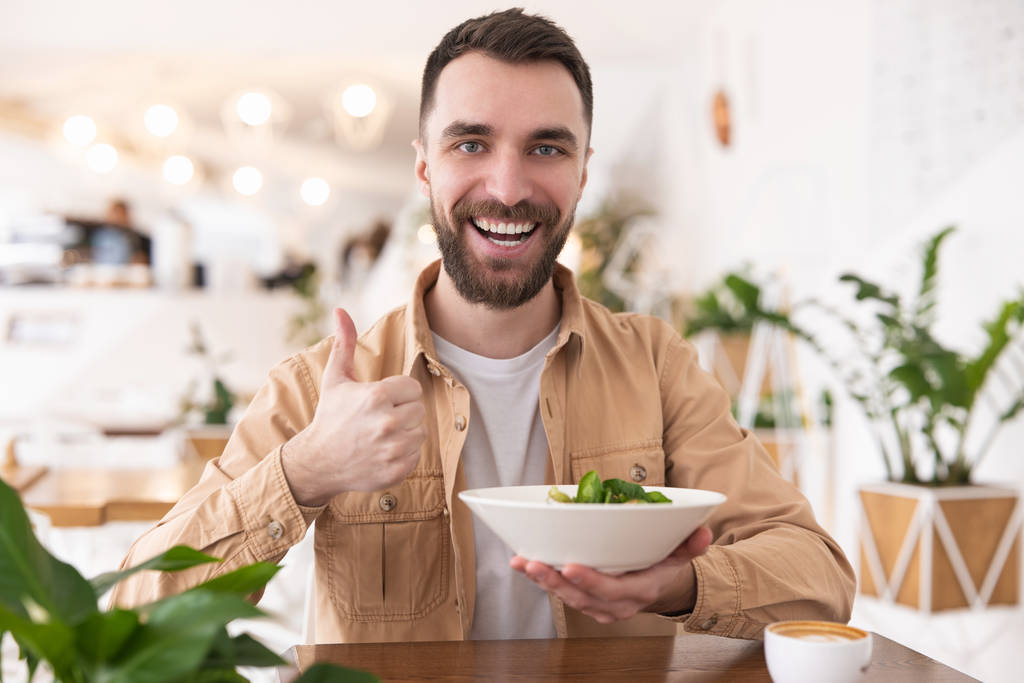 jovem barbudo homem segurando prato de salada em uma mão mostra como enquanto come seu almoço no café durante o intervalo, conceito vegan
. - Foto, Imagem
