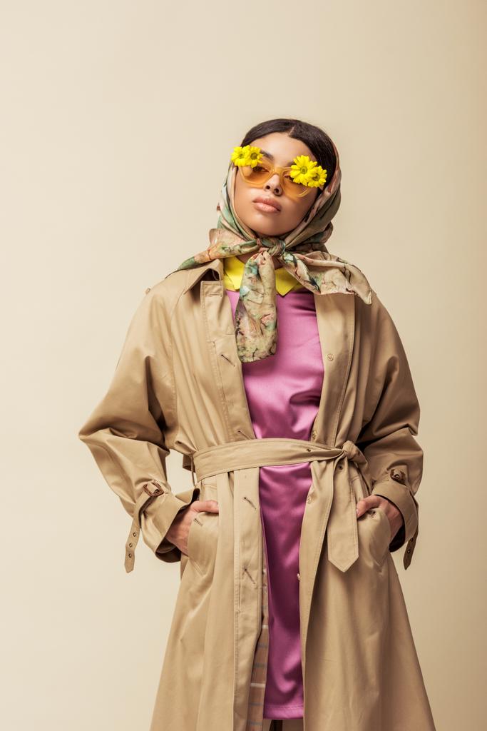 trendy african american girl in sunglasses with flowers, head scarf and trench coat standing with hands in pockets isolated on beige  - Photo, Image