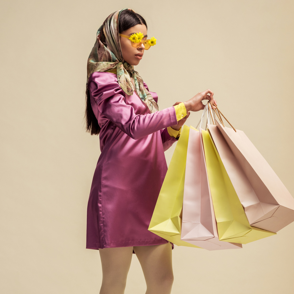 trendiges afrikanisch-amerikanisches Mädchen in Sonnenbrille mit Blumen, Kopftuch und Kleid mit Einkaufstaschen in Beige  - Foto, Bild